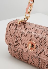 Petit sac à main rose imprimé serpent avec fermeture à fermeture tournante dorée et anse chaîne épaisse rose et dorée sur une surface blanche.
