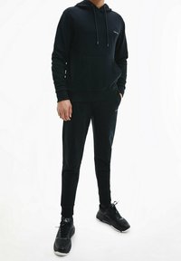 Sudadera negra con bolsillo frontal y cordones, combinada con joggers ajustados a juego, ambos hechos de tejido suave. Estilizada con zapatillas negras.