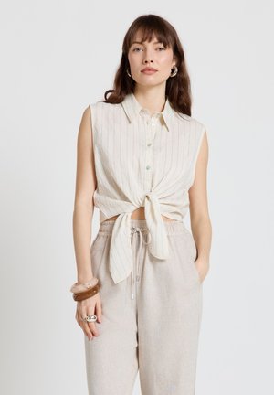 Femme portant un haut beige sans manches boutonné et noué à la taille, avec un pantalon beige clair à cordon, accessoirisée de bracelets et de bagues.