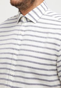 Camicia a righe bianca e azzurro chiaro; presenta un colletto all'inglese e un tessuto testurizzato con vestibilità rilassata.