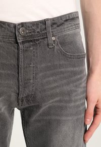 Harmaat farkut, joissa on kapea malli, viisi taskua ja metallinen napitus. Materiaalissa on hieno rakenne ja hienovarainen haalistuminen.