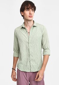 Camisa de manga larga verde claro con un patrón texturizado, cierre de botones en la parte delantera y mangas remangadas, combinada con pantalones morados claros.