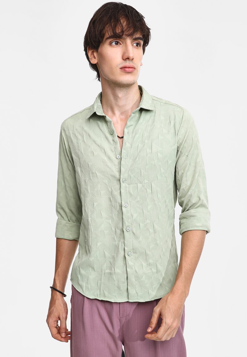 Camisa de manga larga verde claro con un patrón texturizado, cierre de botones en la parte delantera y mangas remangadas, combinada con pantalones morados claros.