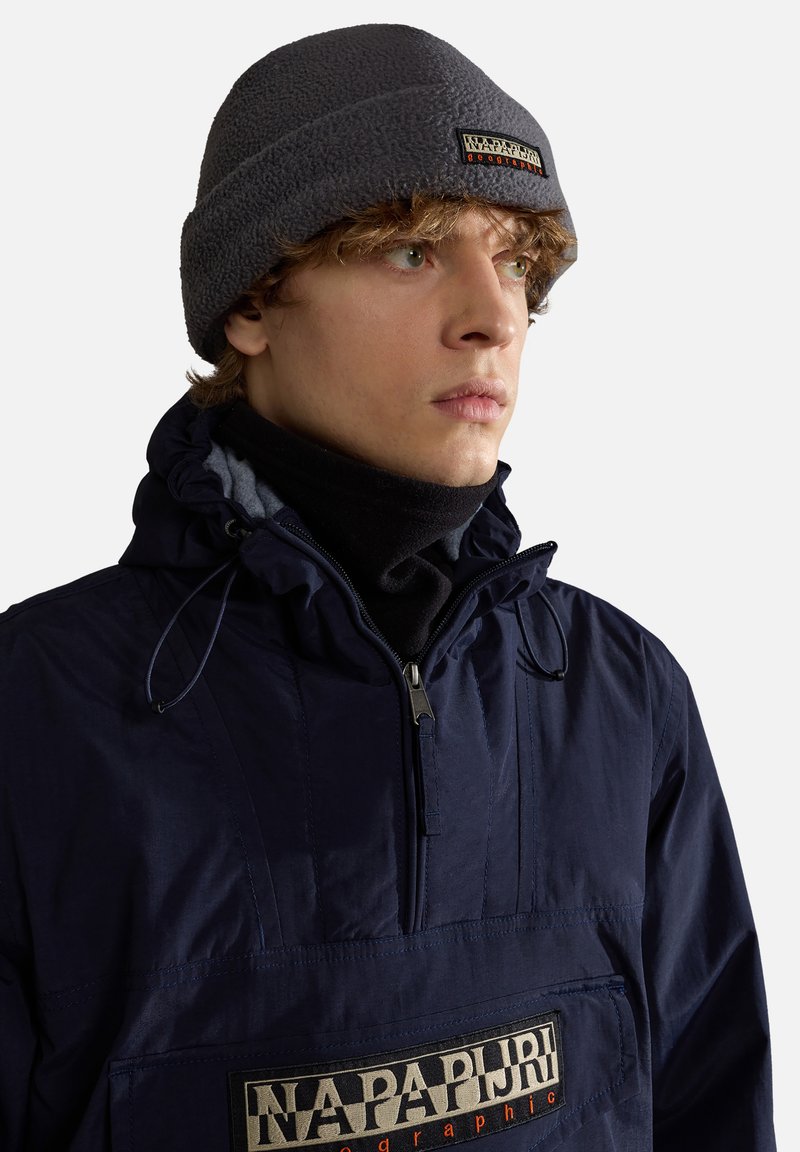 Chaqueta de pullover azul marino con cuello ajustable, cierre frontal con cremallera y un destacado parche de logo en beige y negro. Lleva un gorro de forro polar gris en la cabeza.