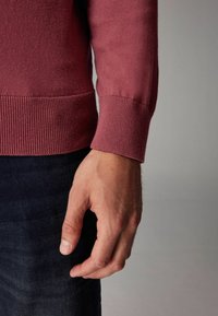Bordeauxfarvet sweater med ribbet kant og manchetter. Stoffet ser blødt og tekstureret ud og er kombineret med mørk denimjeans. Hånden hviler ved siden.