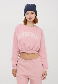 Felpa cropped rosa con la scritta "PHOENIX" in lettere crema, abbinata a pantaloni della tuta rosa coordinati. Il modello indossa un cappellino blu navy.