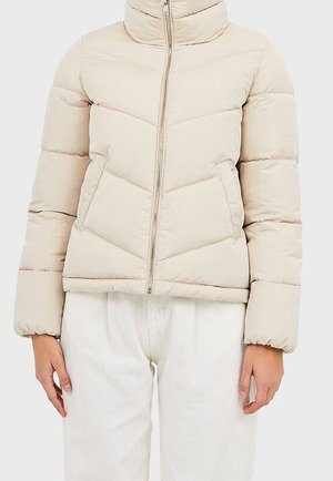 Veste matelassée beige avec fermeture éclair frontale et poches latérales, portée sur un pantalon blanc texturé.