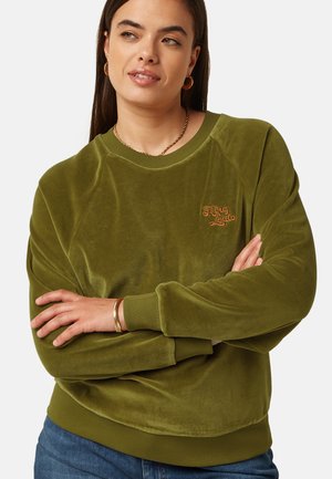 Kvinde med langt, mørkt hår iført guldringøreringe, en guldkæde og en olivengrøn sweatshirt med armene krydset.