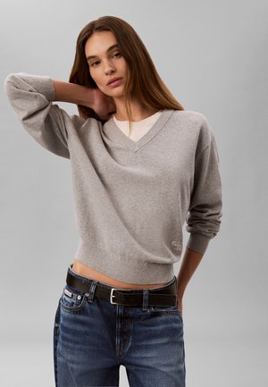 Femme aux longs cheveux bruns portant un pull gris à col en V et un jean bleu avec une ceinture noire sur un fond gris uni.