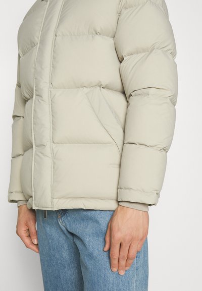 Lys beige puffjakke med quiltede sektioner, lynlåsåbning foran og sidelommer. Blød tekstur og elastiske manchetter, parret med denimjeans.