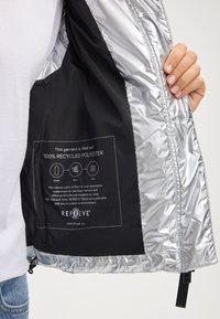 Wnętrze srebrnej kurtki pokazujące etykietę z napisem "100% poliester z recyklingu" na czarnym tle z instrukcją pielęgnacji.