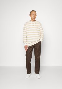 Beżowo-biała bluza w paski z kieszenią z przodu i sznurkami, w połączeniu z brązowymi spodniami i białymi trampkami. Prosty i casualowy design.