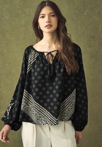Blouse noire à manches longues, avec nœud à la cou et franges. Présente des motifs blancs complexes et une coupe ample, fabriquée en tissu léger.