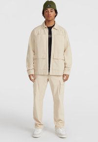 Beige corduroy zipjack met twee voorzakken, gedragen over een zwart shirt. Beige cargobroek met meerdere zijzakken. Witte sneakers.