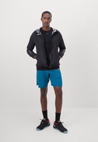 Sort letvægtsjakke med grå hætte, kortærmet sort skjorte, teal shorts og sorte sneakers med røde detaljer, stående på en neutral baggrund.