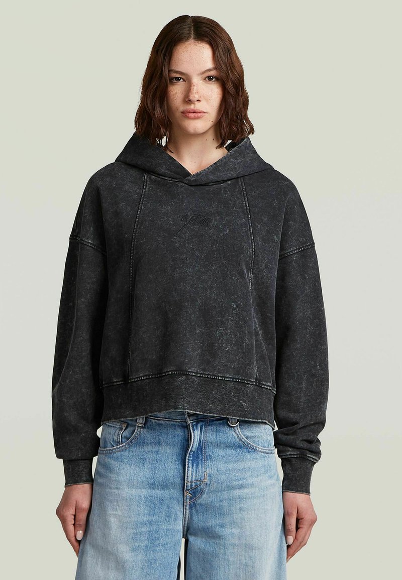 Giovane donna con capelli castani mossi, che indossa un grande hoodie nero scolorito e jeans a vita alta blu chiaro, su uno sfondo chiaro.