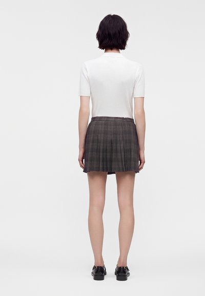 Femme debout, dos tourné, vêtue d’un haut blanc à manches courtes, d’une jupe plissée grise et de mocassins noirs sur un fond blanc uni.