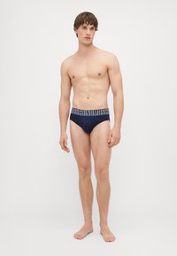 Calções de banho azul-marinho com cintura de logótipo branco, design ajustado e textura suave. Modelo em pé descalço, mostrando o ajuste e o estilo.