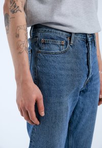 Vaqueros de mezclilla en azul medio con corte recto, que presentan remaches de cobre, costuras en contraste y bolsillos laterales. Textura de tela suave.