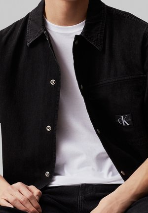 Persona que lleva una chaqueta vaquera negra con botones a presión plateados sobre una camiseta blanca, mostrando un parche visible con el logo de Calvin Klein Jeans.