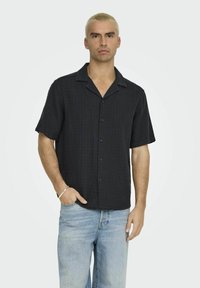 Schwarz kurzärmliges Hemd mit Knopfleiste und strukturiertem Muster, sowie einem eingeschnittenen Kragen, kombiniert mit hellblauen Jeans. Dezente blaue Knöpfe.