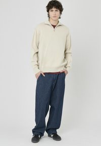 Pull beige en tricot avec col zippé à moitié, associé à un pantalon large bleu à rayures et des baskets noires avec des accents blancs.