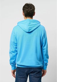 Světlé modré mikina z měkké látky s kapucí na stažení a žebrovanými manžetami, doplněná tmavými džínami. Bez viditelných vzorů nebo log.