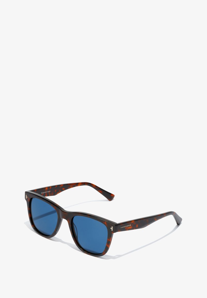 Gafas de sol de carey con lentes azules, montura cuadrada y una textura de plástico suave. Presentan un logo sutil en la patilla.