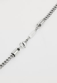 Collier en chaîne en métal argenté avec un fermoir mousqueton et un design de maillons ovales plats, présentant une finition lisse et un détail de logo gravé.