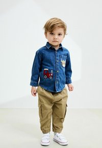 Camicia di jeans con patch a forma di trattore, abbinata a pantaloni cargo beige e sneakers bianche. La camicia ha maniche lunghe e una tasca sul petto.