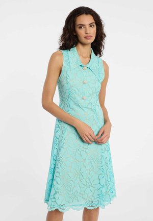 Femme aux cheveux bouclés foncés portant une robe d'été sans manches en dentelle florale bleu clair avec grand col et boutons en perle, debout devant un fond blanc.