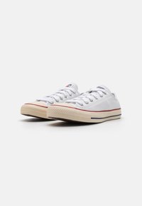 White canvas sneakers with round toes, lace-up design, contrasting red stripe, and beige rubber soles featuring textured patterns.