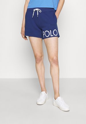 Osoba nosząca niebieskie spodenki ze białym sznurkiem i dużym białym napisem "POLO" na udzie, zestawione z białymi sneakersami i niebieską koszulą.