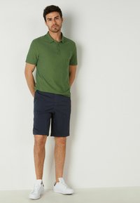 Grön kortärmad polo, marinblå shorts och vita sneakers. Polon har en subtil textur och en treknappsgatering.