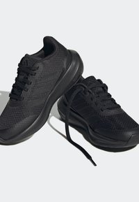 Des chaussures de sport noires présentent un dessus en tricot texturé, une semelle en caoutchouc lisse, un col rembourré et des lacets plats. Deux bandes sur les côtés sont bien visibles.