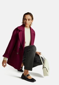 Abrigo de lana magenta con grandes solapas y dobles botones, combinado con un suéter crema y pantalones grises. Zapatos negros con detalles de hebilla.