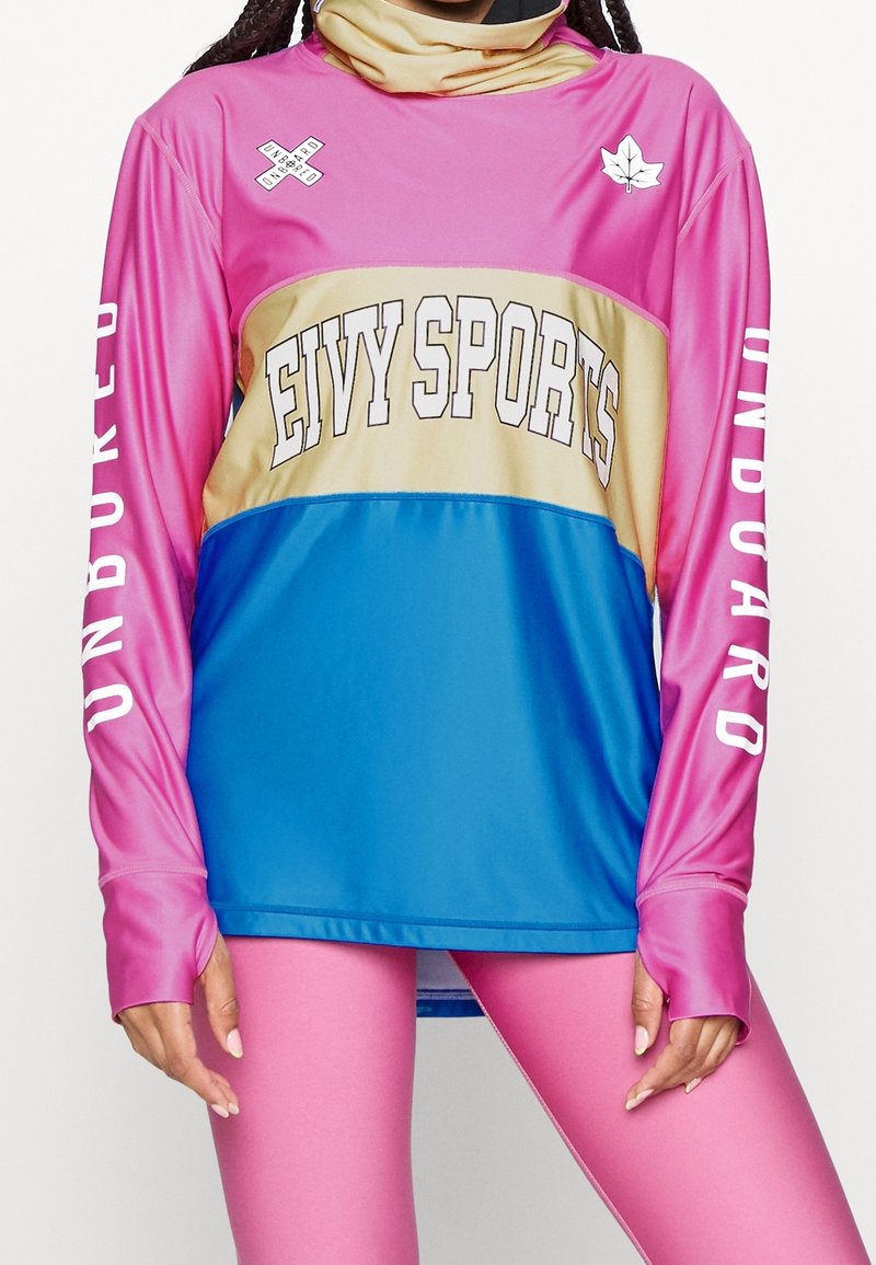Personne portant un t-shirt de sport en blocs de couleur rose, or et bleu avec les inscriptions "Eivy Sports" et "Unboard", associé à des leggings roses.