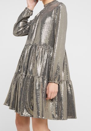 Frau, die ein langärmeliges, goldfarbenes, metallisch glitzerndes Kleid mit gestuftem Rock und hohem Kragen trägt, zu sehen von der Schulter bis zur Mitte des Oberschenkels.