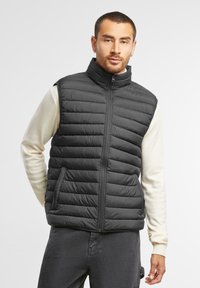 Schwarze gepolsterte Weste mit vertikal gestepptem Muster, hohem Kragen und vorderem Reißverschluss, getragen über einem hellen Langarmshirt.