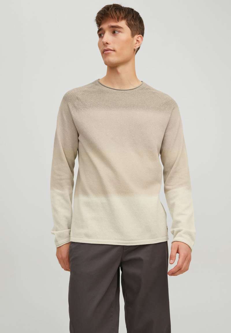 Un pull à col rond à manches longues beige avec un motif texturé, une couleur dégradée allant du clair au foncé, et des poignets côtelés. Associé à un pantalon sombre.