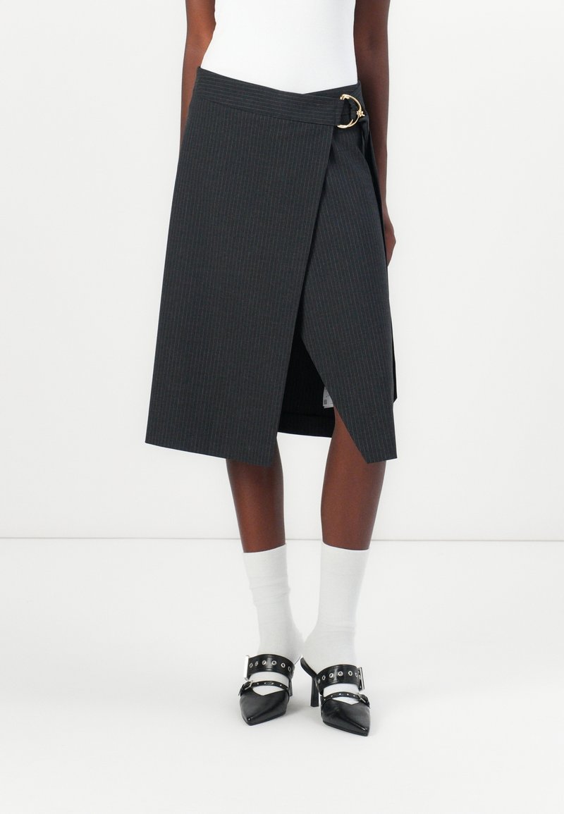 Jupe portefeuille noire à fines rayures longueur genou avec boucle latérale, portée avec des chaussettes blanches et des talons noirs pointus.