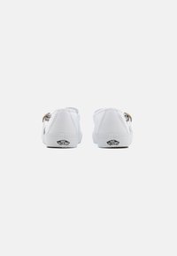 Zapatillas de lona blancas con suelas planas, que cuentan con un acento dorado en el talón y un logotipo negro en la suela. Diseño de punta redondeada.