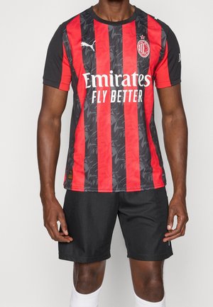 Mężczyzna ubrany w koszulkę piłkarską AC Milan z czerwono-czarnymi pionowymi paskami, logo Emirates Fly Better, czarne spodenki i białe skarpetki.