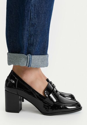 Mocasines negros brillantes con tacón de bloque, usados con vaqueros azul oscuro remangados, destacando el acabado pulido del zapato y el diseño cuadrado de la puntera.
