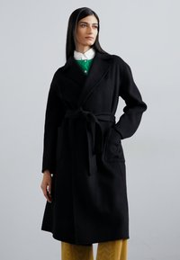 Zwarte, lange jas met een ceintuur in de taille, zijzakken en brede revers. Gladde, textuurstof. Gestyled over een groene trui met een witte kraag.