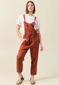 Rostfärgade hängselbyxor med justerbara remmar, knappdetaljer och knytmidja. I kombination med en vit t-shirt och metalliska skor.