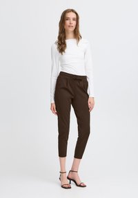 Naine, seljas pruunid pahklaiuse püksid, millel on tõmbluku talje ja valge pikkade varrukatega topp, seisab paljajalu tavalise valge taustaga.