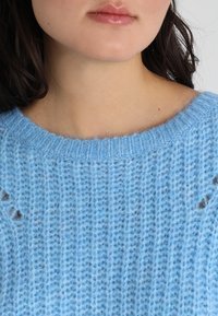 Ljusblå stickad tröja med rund halsringning, som har ett strukturerat mönster och små, mörkgrå detaljer på axlarna.