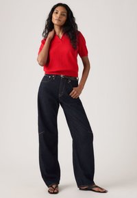 Κόκκινη πόλο μπλούζα με κοντά μανίκια συνδυασμένη με σκούρο μπλε παντελόνι με φαρδιά μπατζάκια και κίτρινα ραφτικά. Το μοντέλο φοράει μαύρα σαγιονάρες.