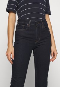 Pantalones vaqueros de mezclilla azul oscuro con cinco bolsillos, costuras amarillas, cierre de botón y cremallera, y un diseño ajustado.